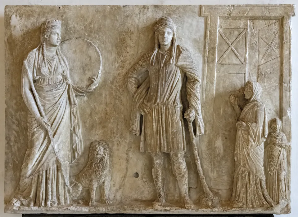 A white marble relief: at left the goddess Cybele holds a frame drum, a lion at her feet; in the centre stands the youth Attis in a Phrygian cap and leggings, holding a shepherd's crook; at right a veiled woman and child approach through a doorway.