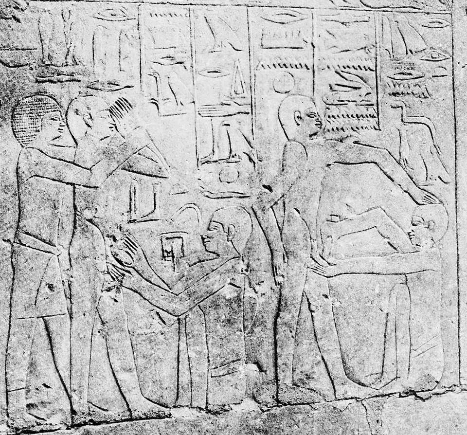 A carved limestone wall relief under a band of hieroglyphs: on the left a kneeling man operates on a standing youth, whose companion holds his arm; on the right another youth is held from behind by an attendant while a kneeling operator works.