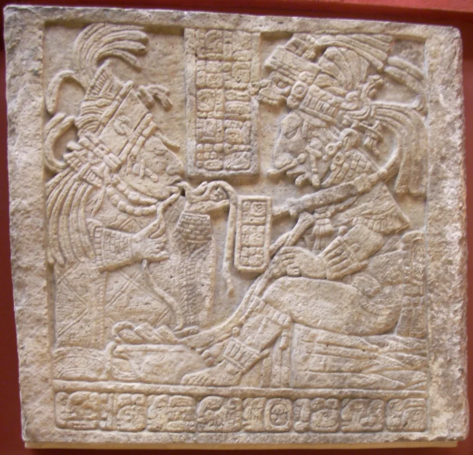 A square carved limestone lintel: at left a kneeling woman in an elaborate headdress pulls a cord through her tongue; at right a seated man in a feathered headdress holds a long pointed perforator; glyph panels between and below.