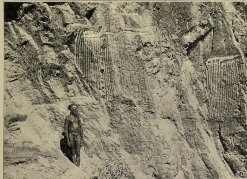 A man stands at the foot of a steep rock face painted with long panels of vertical stripes.