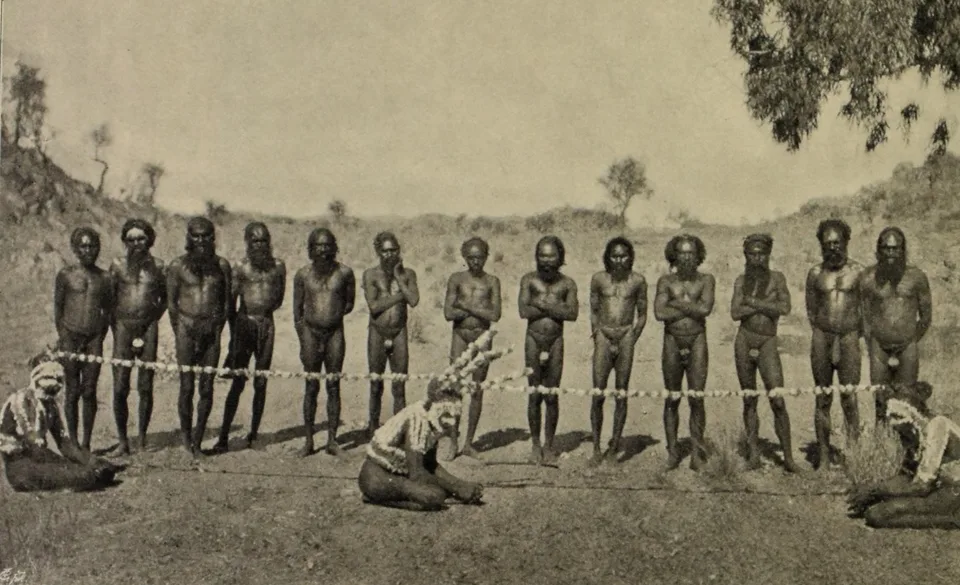 A row of Arrernte men stand behind a long decorated cord stretched across open ground, with three decorated performers seated or kneeling in front of it.