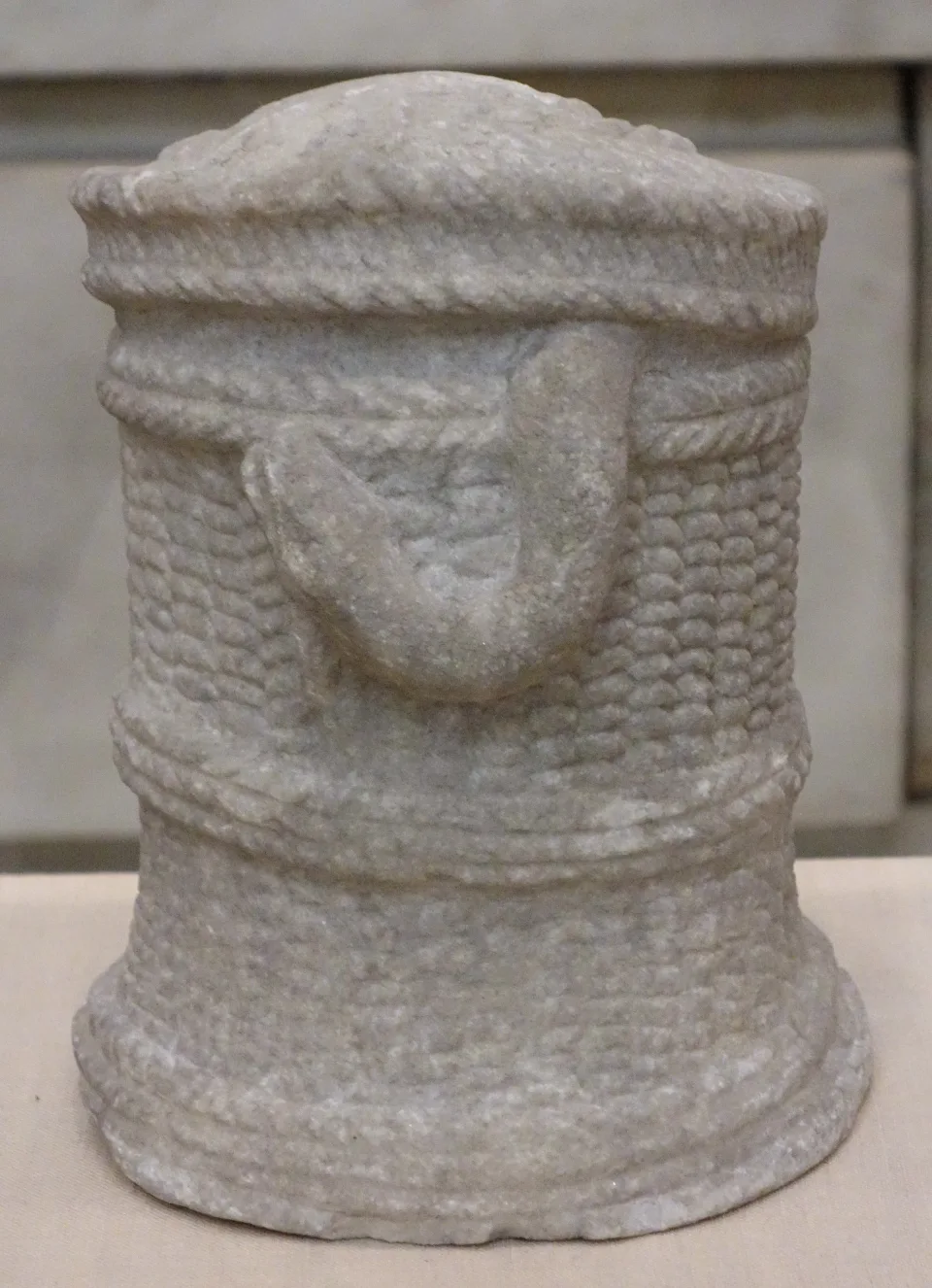A small cylindrical marble basket carved to imitate woven wicker, with a rope-twist rim and a closed lid, and a thick serpent carved in relief looping across its side.