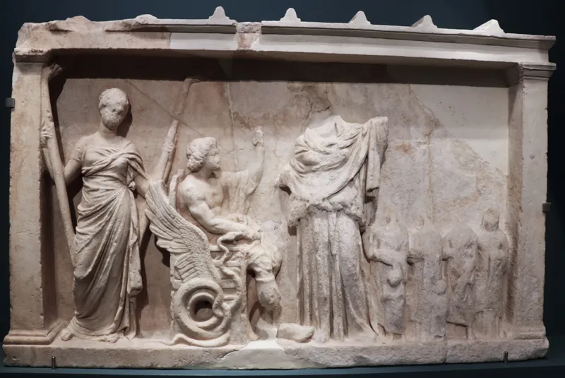 A marble relief: two standing goddesses flank a young man seated on a chariot whose wheel is a coiled serpent; small worshippers approach from the right.
