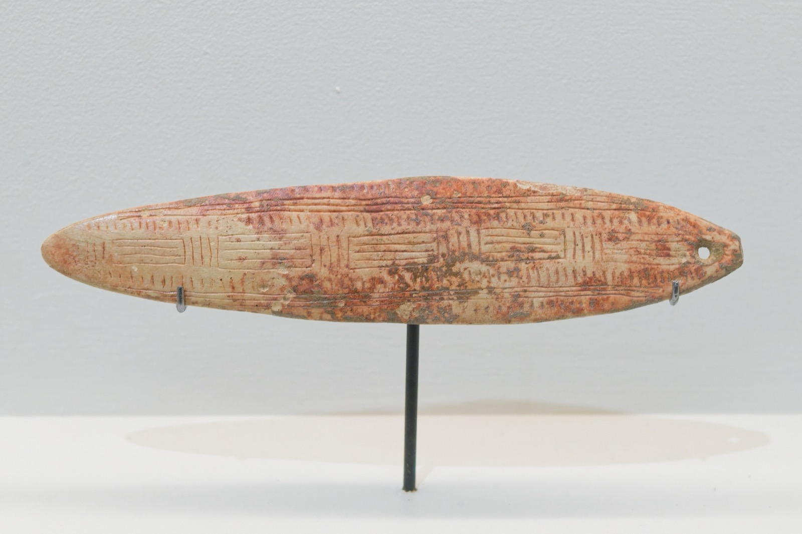 Elongated reindeer-antler blade streaked with red ochre and incised with ranks of short parallel lines, pierced at its narrow end, mounted on a museum stand.