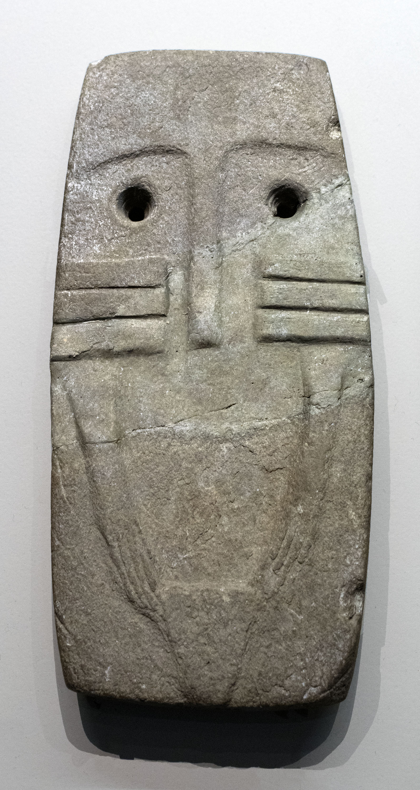 Grey schist plaque carved with two round staring eyes, a long nose ridge and incised whisker lines, above a robed lower body.