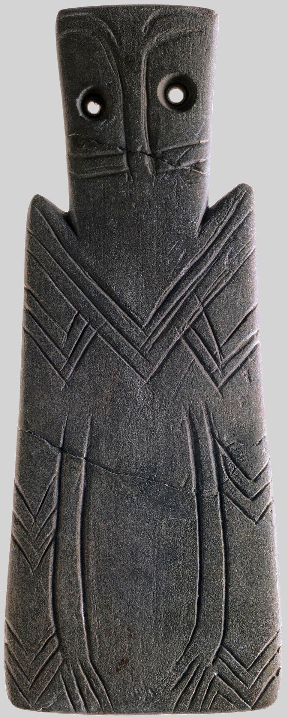 Dark slate plaque with two drilled owl eyes at the top and deep incised chevrons running down its trapezoidal body.