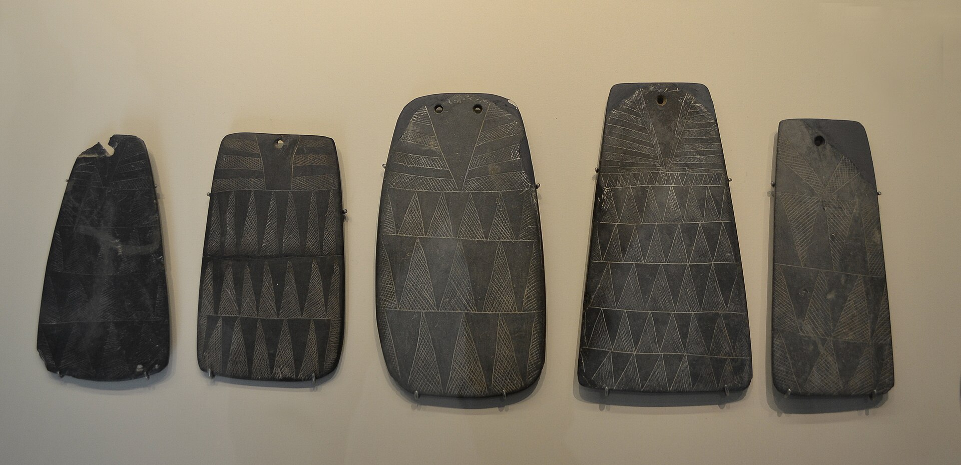 Five dark engraved slate plaques of different widths and heights mounted in a row, each covered in incised triangles and bands, each pierced at the top.