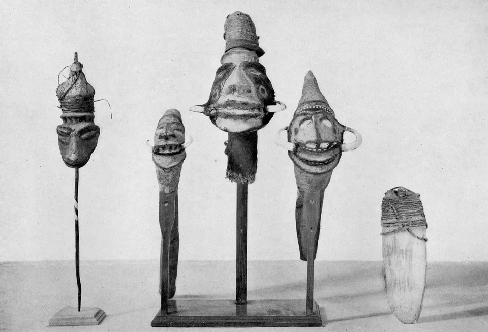Four carved heads with tusked grinning faces mounted on sticks and stands, and at the far right a pale bullroarer bound with cord.
