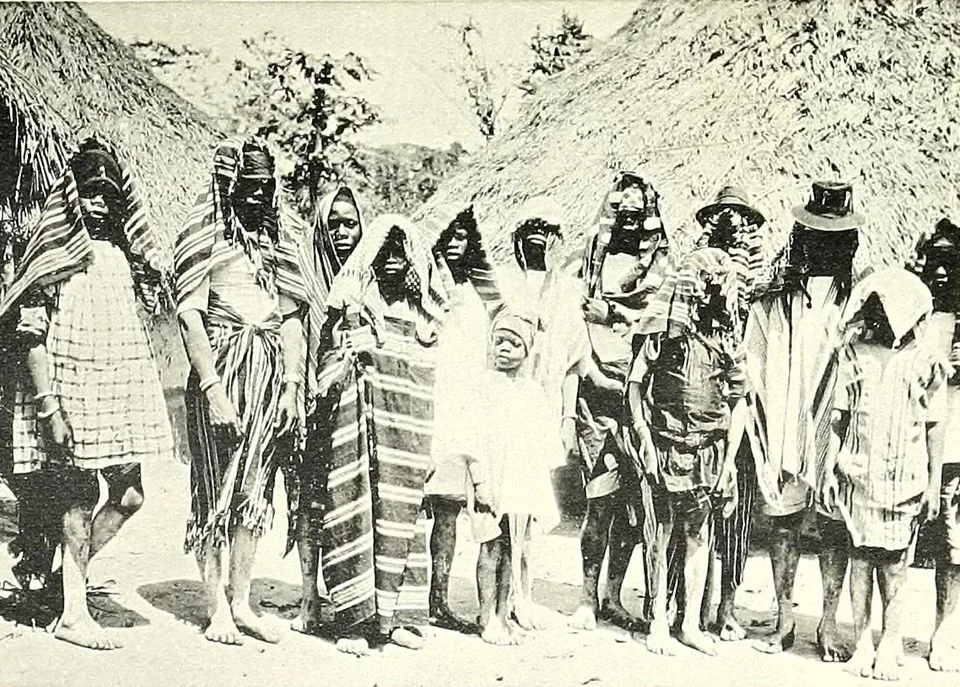 A row of boys and youths wrapped in striped cloths and shawls stand barefoot in front of thatched houses, some with their heads hooded, staring at the camera.