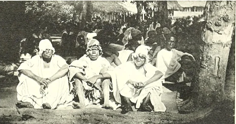 Three young men wrapped in white cloths and head-cloths sit on the ground under a large tree, a crowd behind them.