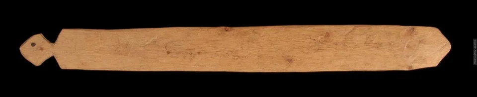 A long pale softwood bullroarer blade tapering to a point, with a diamond-shaped end where the cord is seated.