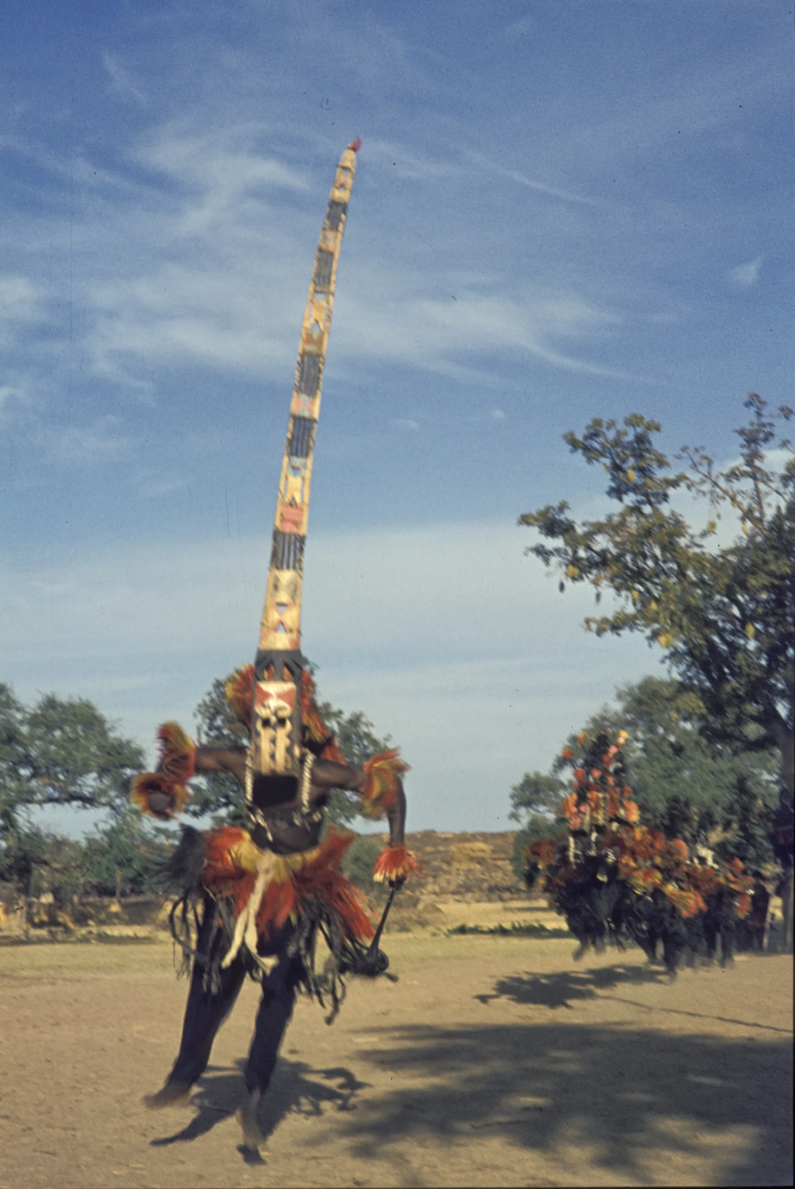 A Dogon dancer in red and yellow fibres wearing a sirige mask, a painted plank several metres tall, against a blue sky.