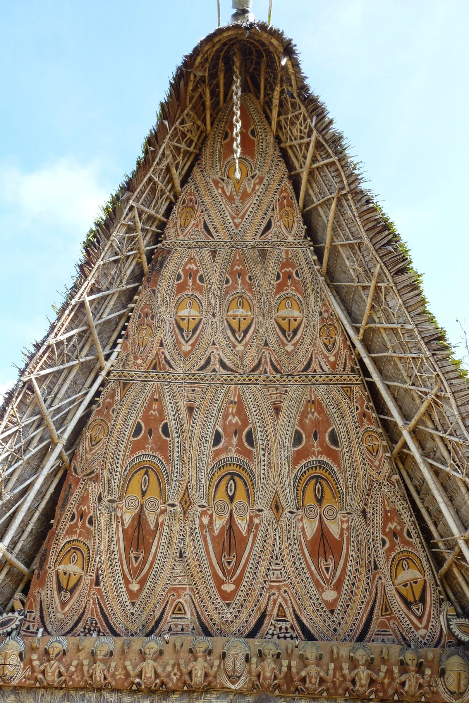 The soaring triangular painted gable of a spirit house seen from below, with tiers of large painted faces in ochre, yellow, black and white.