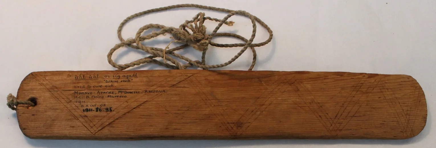 A long, straight-sided wooden blade with a rounded end and a cord through the hole, incised with zigzag triangles and inscribed in ink.