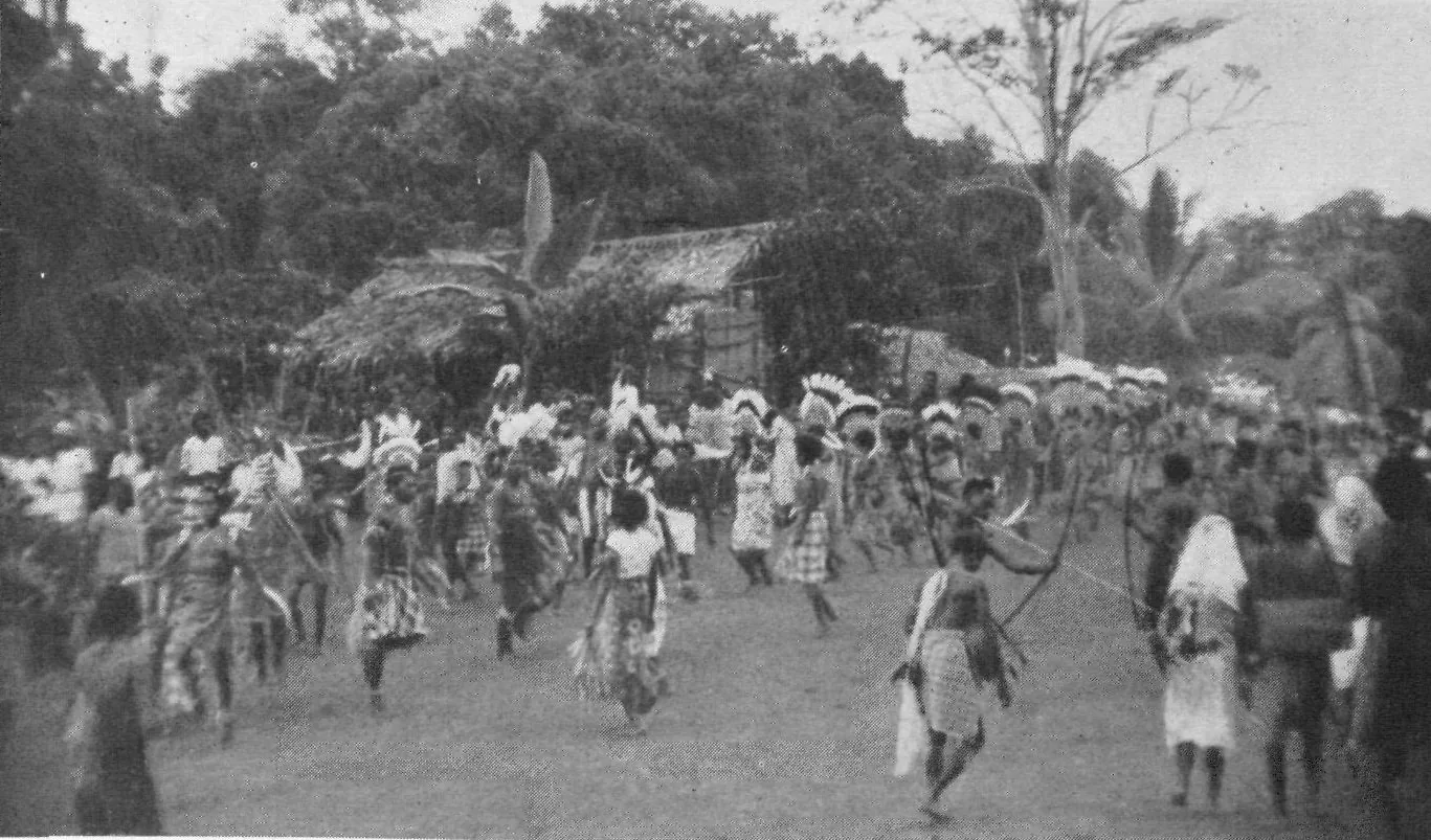 A crowd of dancers in feather headdresses and fibre skirts streams across an open square toward the camera, bowmen at the front, a thatched house behind.