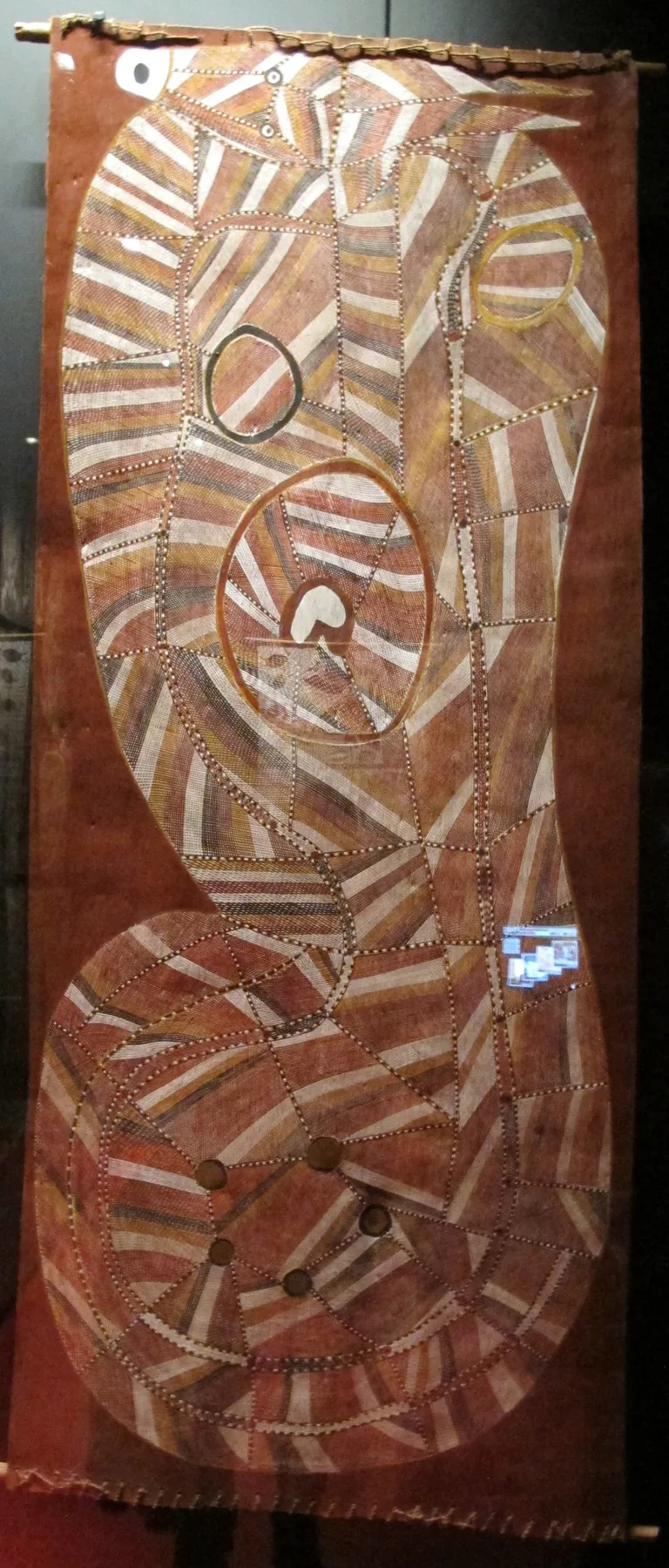 A tall bark painting of a coiled, horned rainbow serpent in fine red and white cross-hatching.