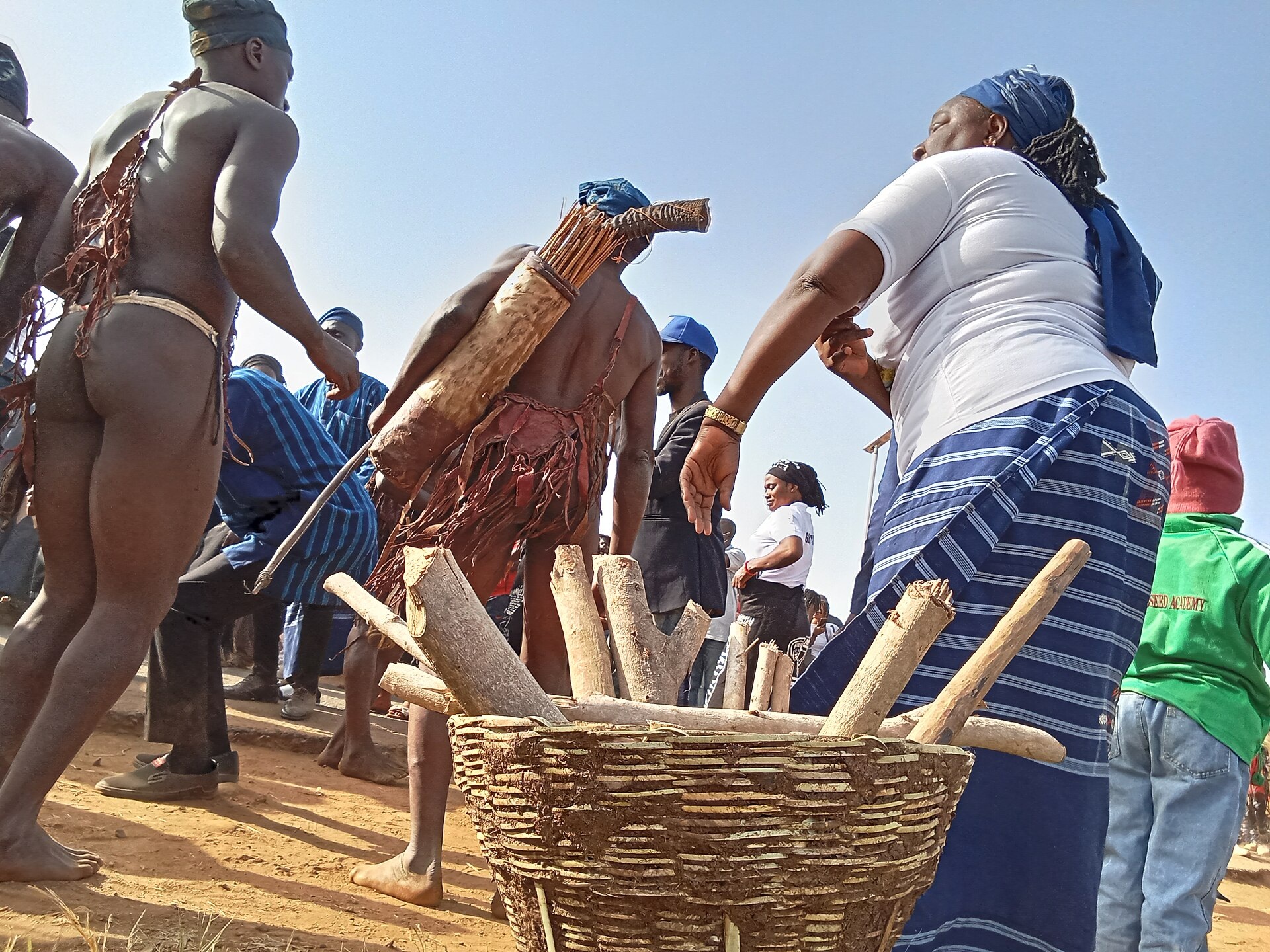 Gbagyi dancers in blue checked cloth and woven costumes performing outdoors around a basket of wooden sticks.