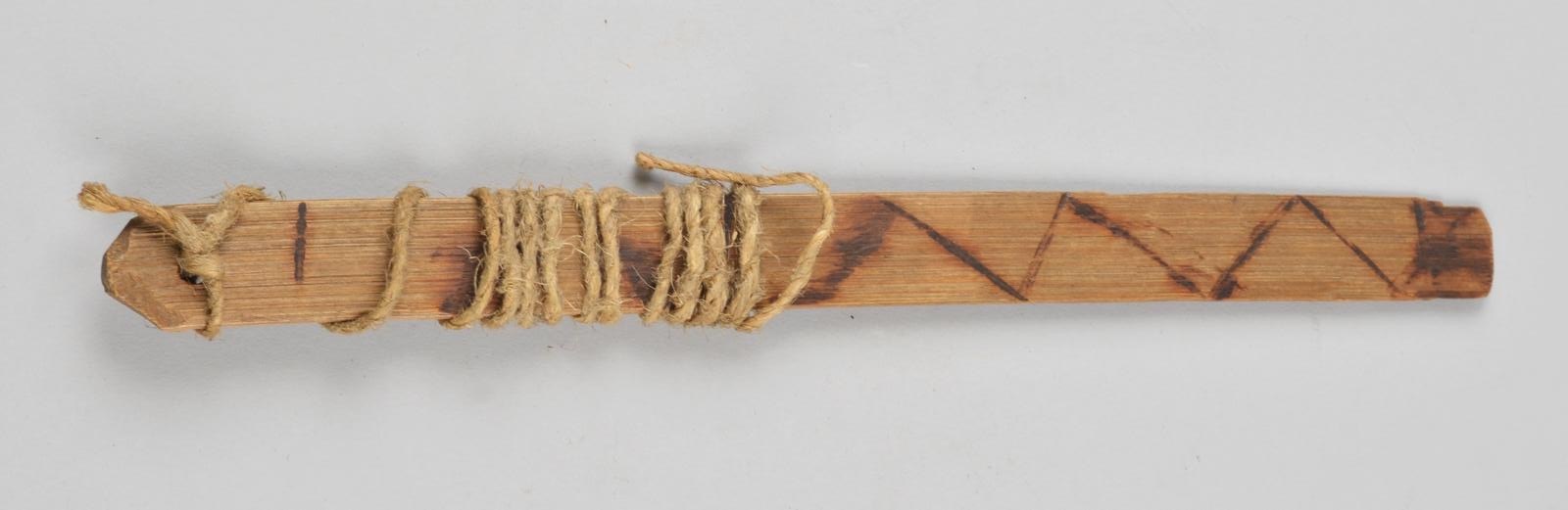 A Gwari bamboo strip with a dark zigzag line burned along much of its length and cord wound around one end.