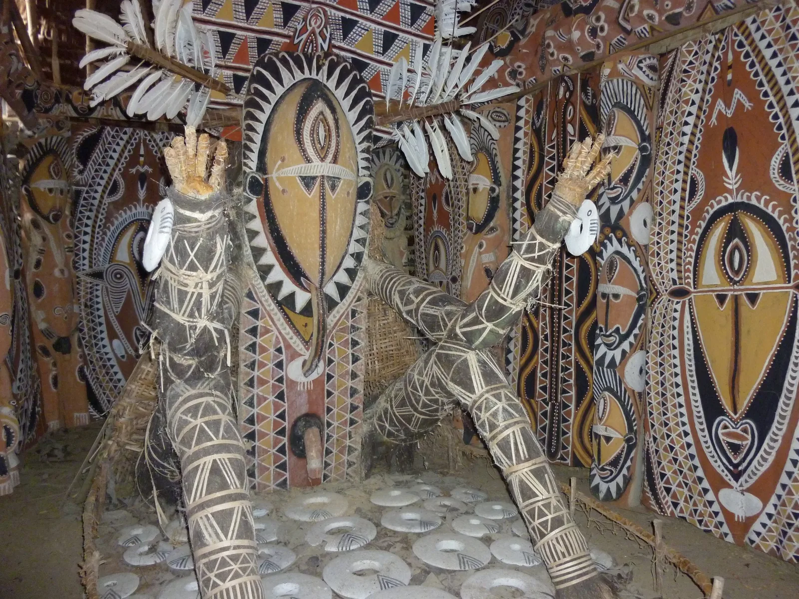 Inside an Abelam spirit house: huge painted ancestral faces in ochre, black and white line the walls, two bound figures with shell rings raise their arms beneath white feather plumes, and the floor is laid with white shell rings.