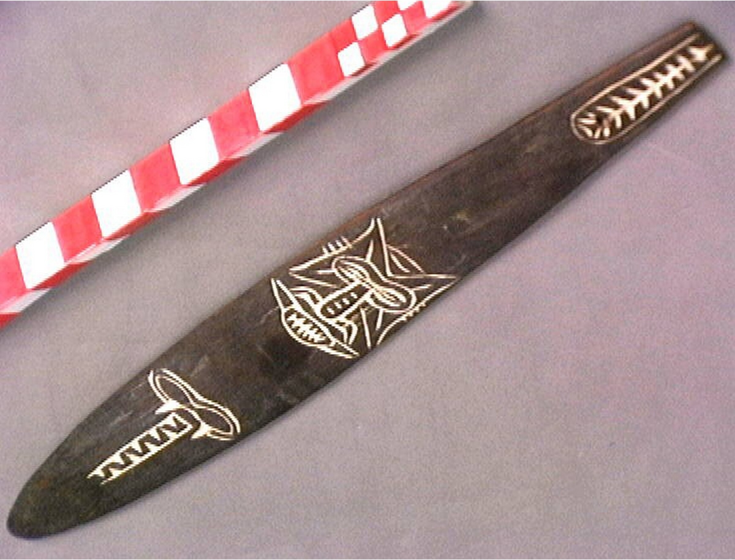 Narrow dark Tami bullroarer with pale lime motifs near both ends and a large face-like design in the middle.