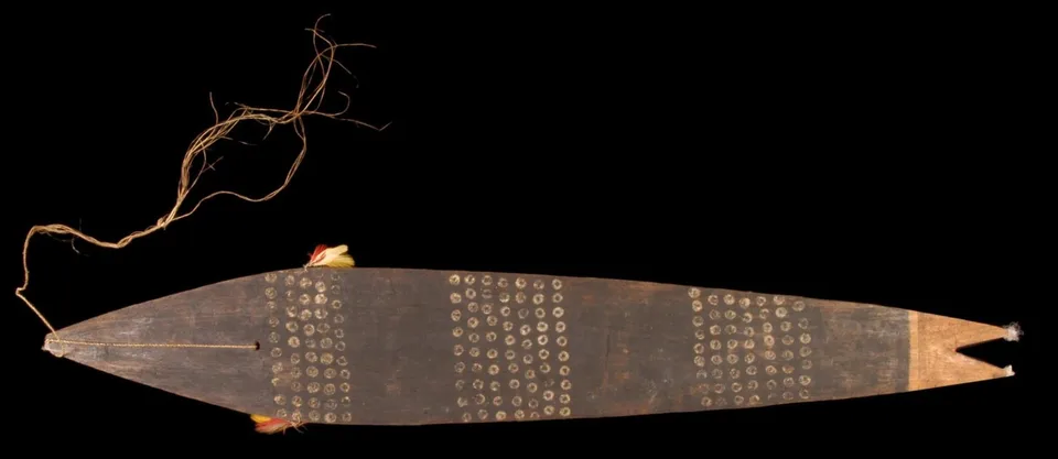 A long dark Bororo bullroarer with a fish-tail notch at one end, painted with three bands of small pale rings, its cord trailing from the pointed end.