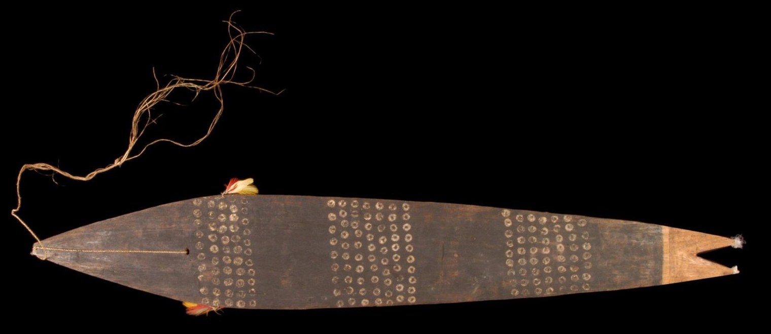A long dark wooden bullroarer photographed against black, decorated with bands of small white dotted circles, with a fibre cord at one end and a notched fish-tail at the other.