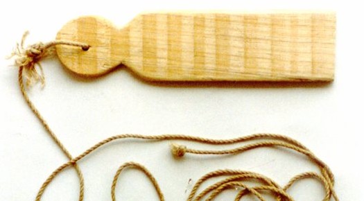 A Basque burruna: a flat pale wooden slat with a round, pierced head and a loose cord.