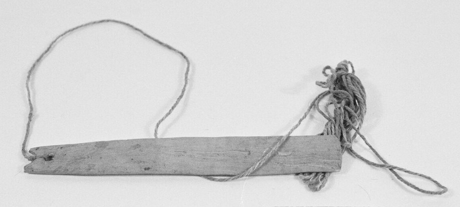 A plain pale wooden bullroarer slat on a looped cord, photographed against a grey background.