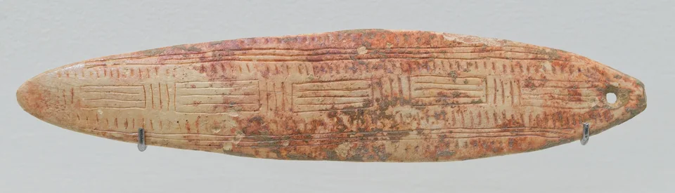 Lens-shaped reindeer-antler bullroarer from La Roche de Birol, stained with red ochre, engraved with panels of parallel lines and pierced at its narrow end.