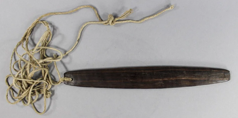 A slender dark wooden blade, tapering at both ends and pierced at one end, tied to a long tangle of pale fibre cord.