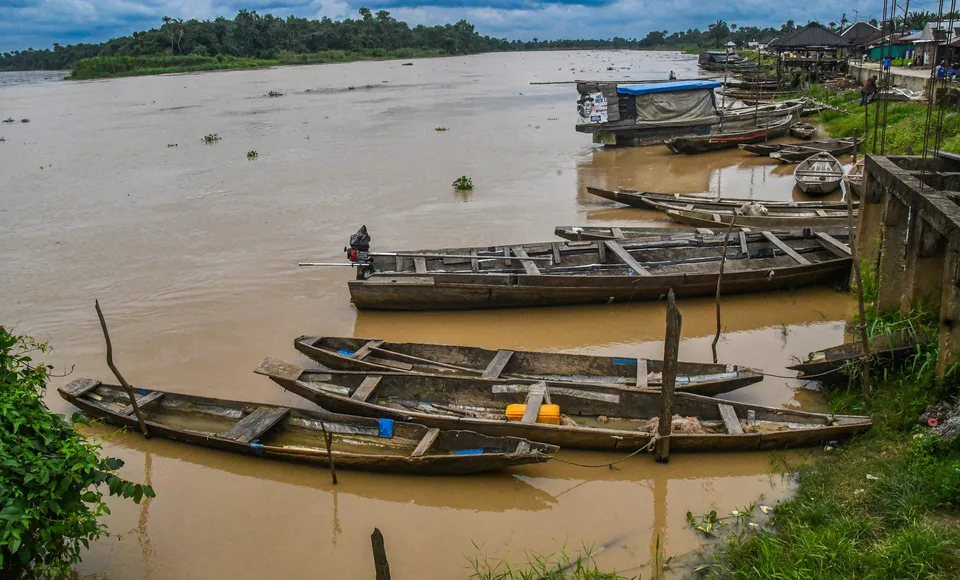Long wooden dugout canoes moored along the muddy bank of a broad brown river; a covered boat and houses on stilts stand upstream, with forest on the far shore.