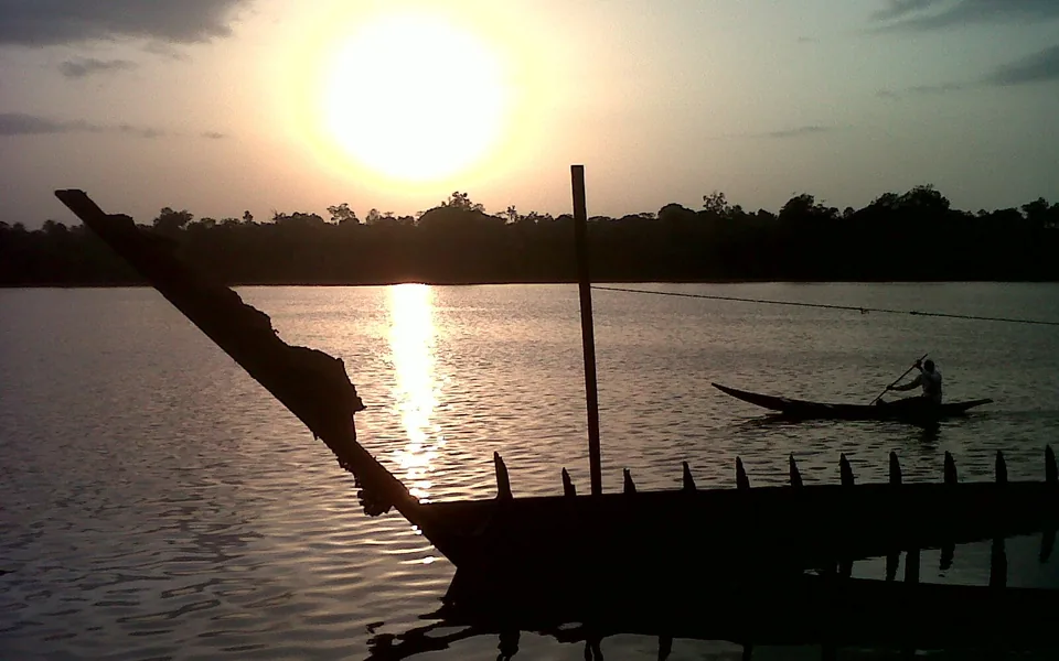 Sunset over a wide creek fringed with dark forest; a man paddles a dugout canoe across the sun's reflection, and the ribs of an old wrecked boat stand black in the foreground.