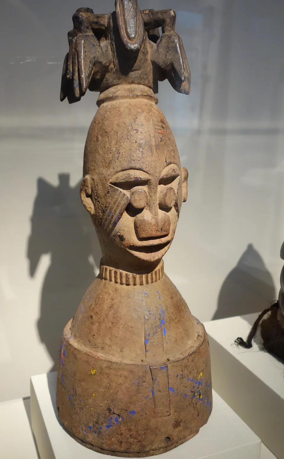 A carved wooden headdress: a human head with scarified cheeks and a ringed neck on a broad bell-shaped base, crowned with carved forms like curving horns.
