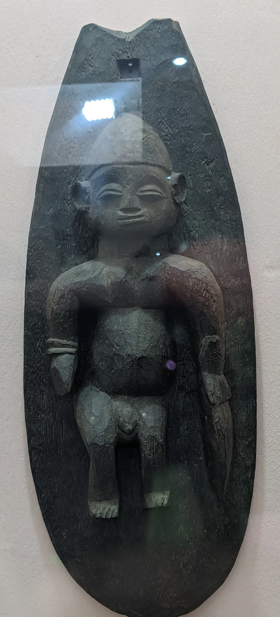 A large dark wooden board, rounded at the bottom and notched at the top, with a hole near the top and a standing human figure carved in relief down its face, displayed behind glass.