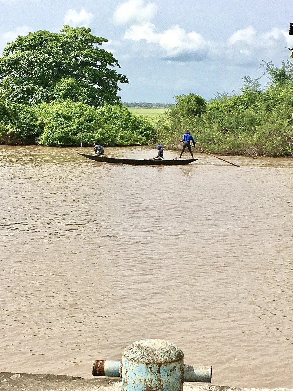 Two men stand in a slim canoe on a wide brown river, one poling, with dense green bush on the near bank and open sky above.