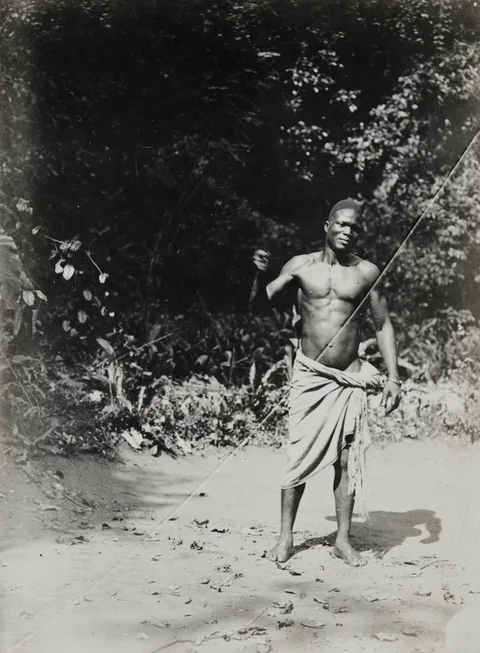 Black-and-white photograph of a young man in a wrapper standing on a sandy forest path, his right arm raised as he whirls a bullroarer; its cord shows as a pale line slanting across the picture.