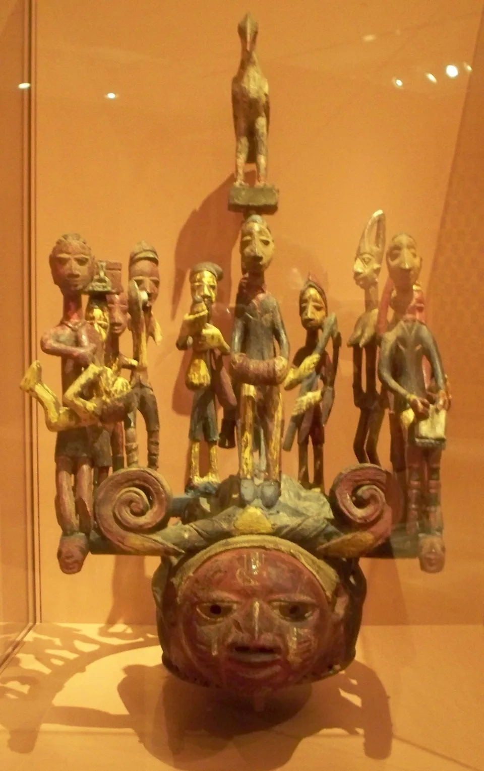 A carved wooden helmet mask with a red-painted face at its base, crowned by a crowded superstructure of small standing figures, a central figure on a tall post above them.