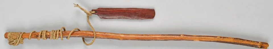 A long, slightly curved wooden stick wrapped with cord at one end, joined by a loop of string to a short reddish wooden blade with bevelled edges.