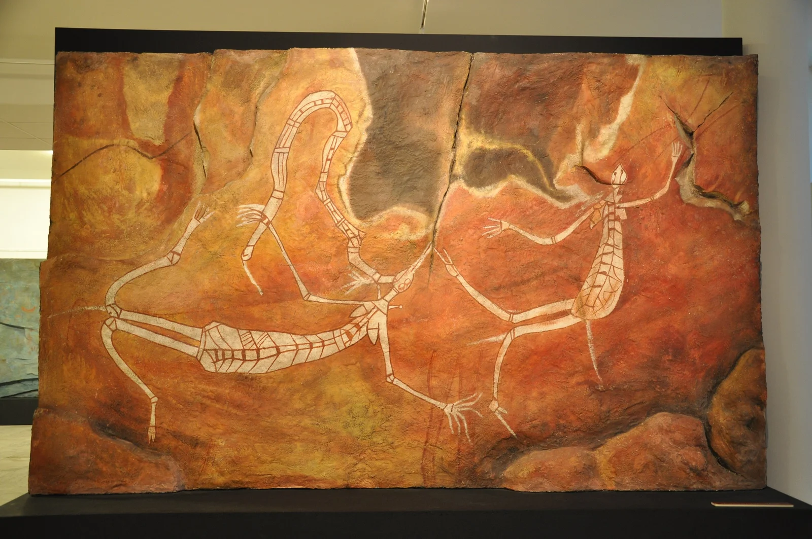 The pale, patterned serpent Burlung loops across a red rock surface beside long-limbed Namaroto spirits, in a museum replica.