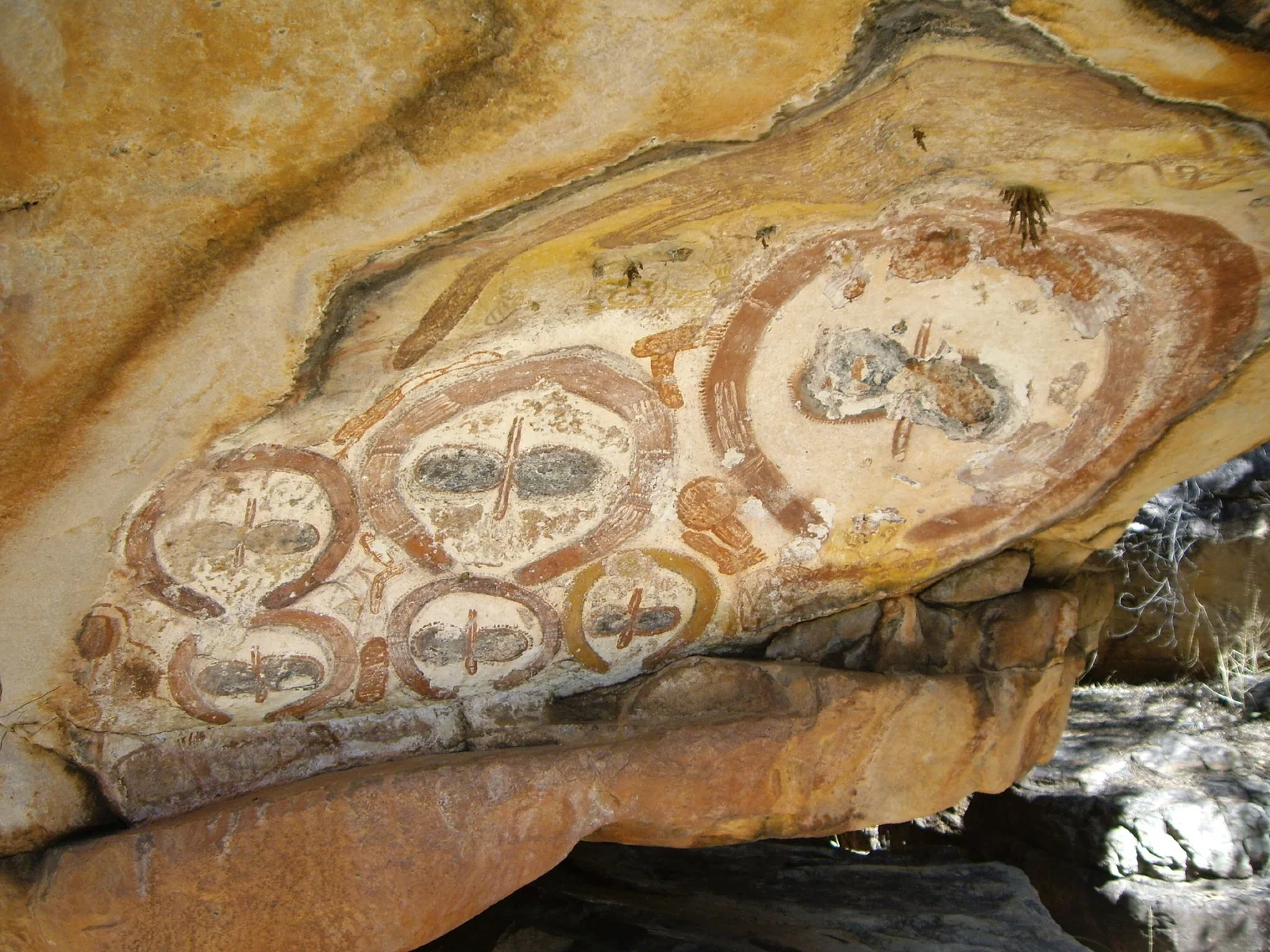 Pale Wanjina figures with large dark eyes and red head surrounds painted beneath a Kimberley rock overhang.