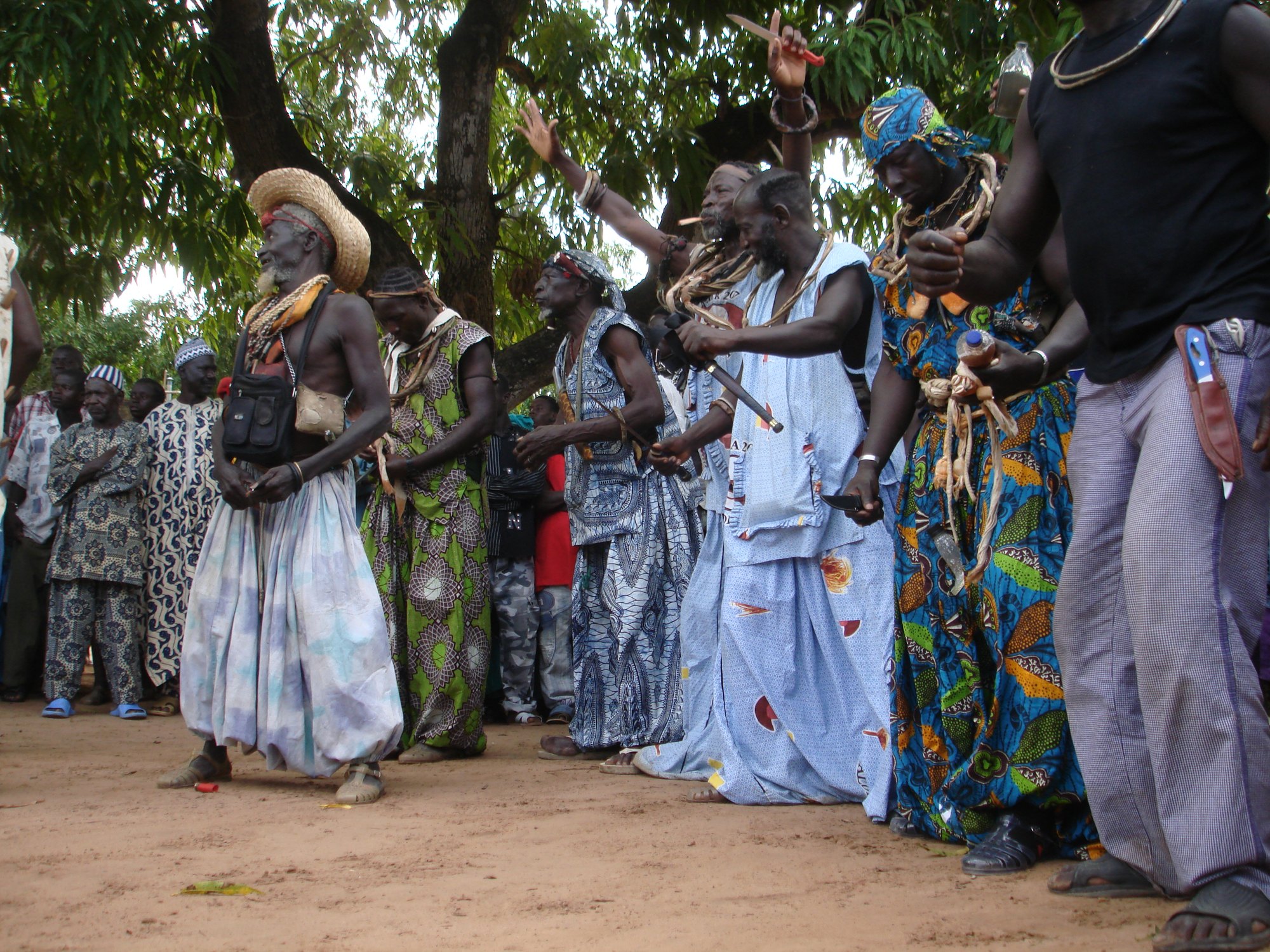 Older Jóola men in hats and long robes dance in a line on red earth under trees, watched by a crowd of men and women.