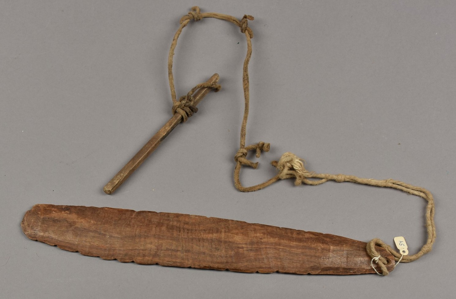 A long brown wooden bullroarer blade with a scalloped edge, tied by a knotted cord to a short wooden handle.