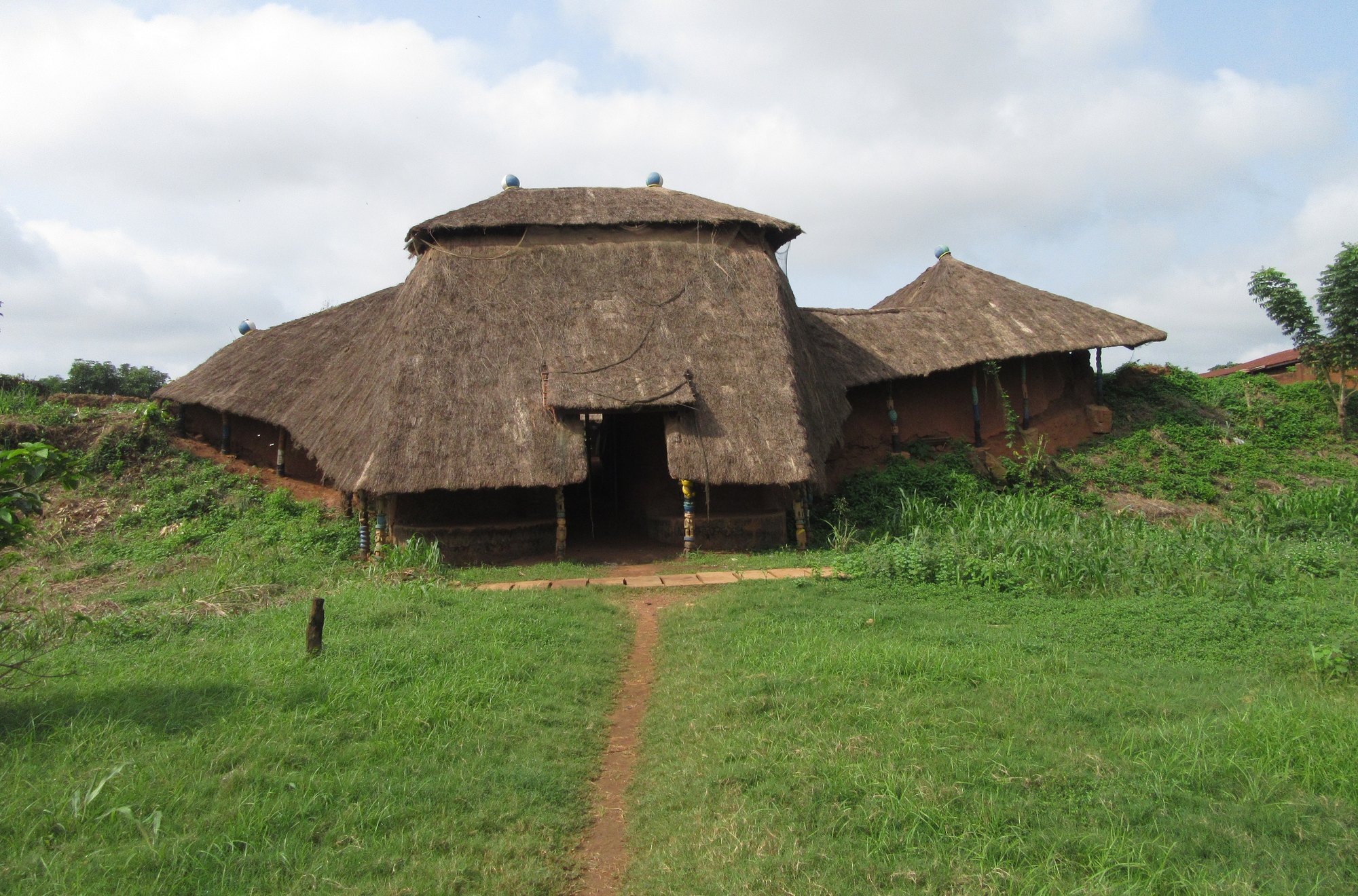 A large thatched gatehouse with a deep doorway, flanked by lower thatched buildings, at the end of a dirt path across a green lawn.