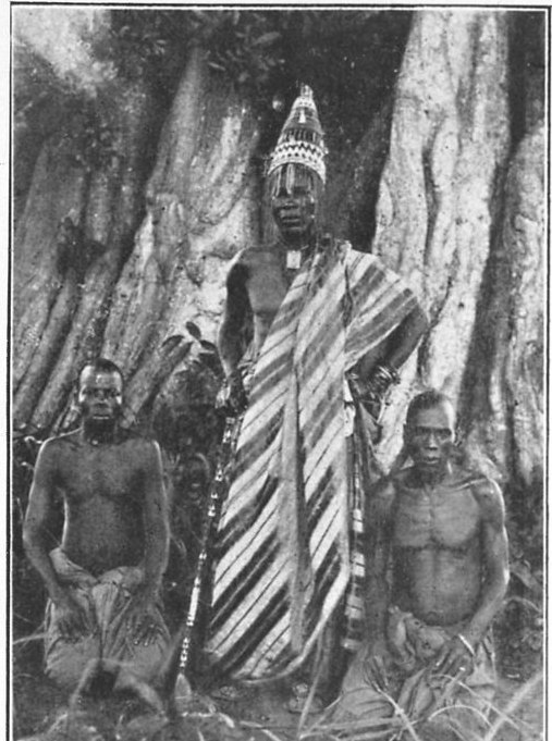 Black-and-white photograph of a tall man in a beaded conical crown and a striped wrapper, standing before huge tree buttresses, with two kneeling men on either side.