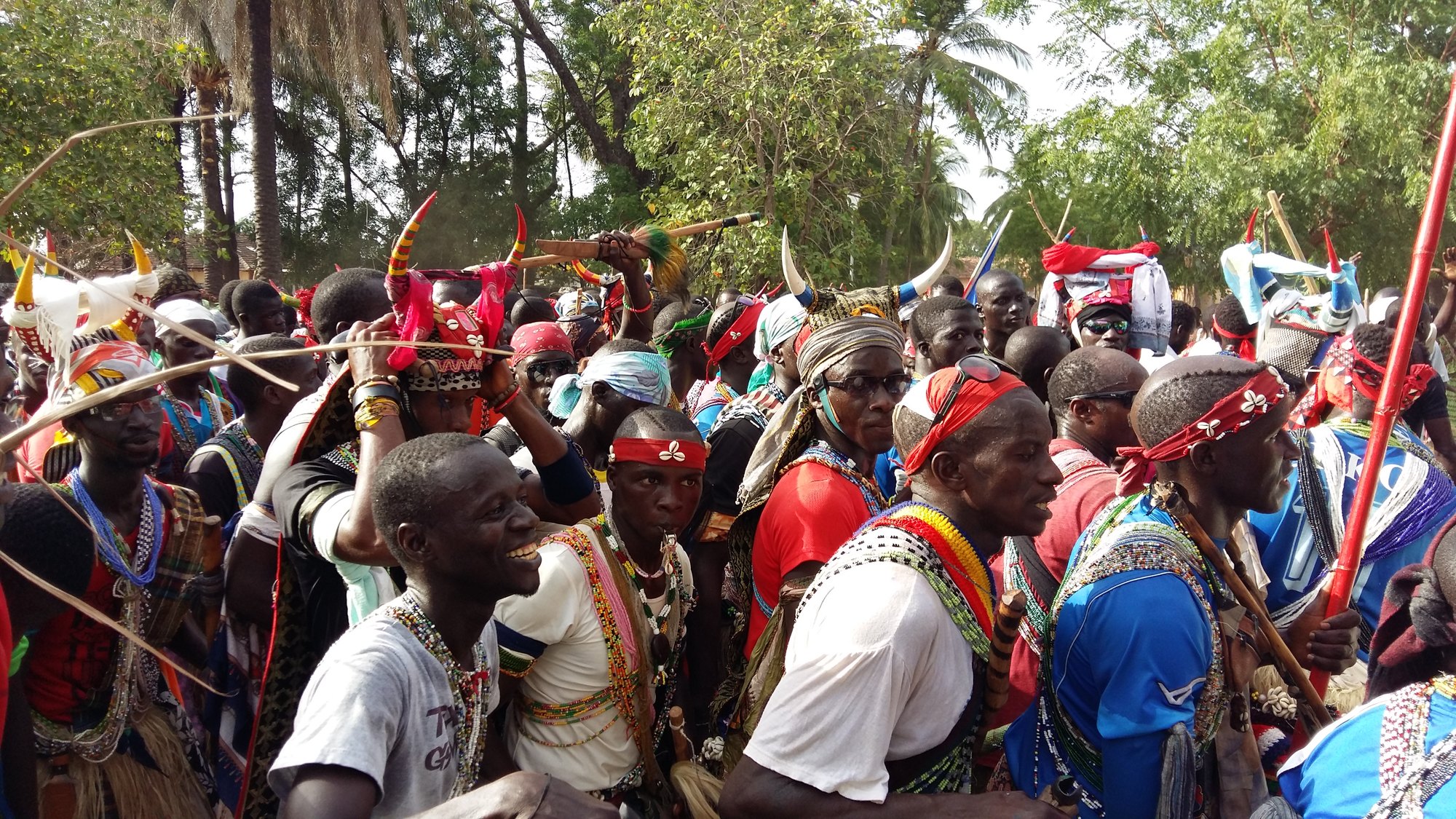 A dense crowd of Jóola men in red headbands, bead sashes and horned headdresses, some carrying sticks and red staffs, gathered under palms and tall trees.