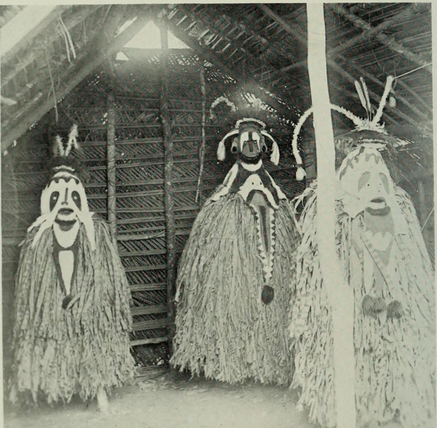 Three tall masked figures in long fringed grass costumes standing inside a thatched house; their faces are painted white with dark eyes and mouths, and feathers rise from their heads.