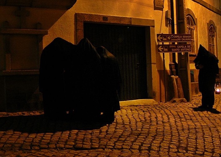 Hooded figures in dark cloaks stand at a cobbled village corner at night, one holding a lantern, beneath signs pointing to two chapels.