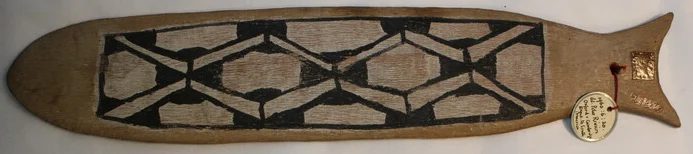 A fish-shaped wooden Kamaiurá bullroarer with a forked tail, its middle painted with a bold black-and-white lattice of hexagons and diagonals.