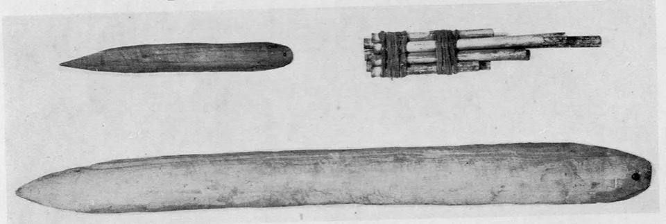 Two plain wooden bullroarers photographed in a museum plate, a long blade below and a short pointed one above, with a bundle of panpipes.