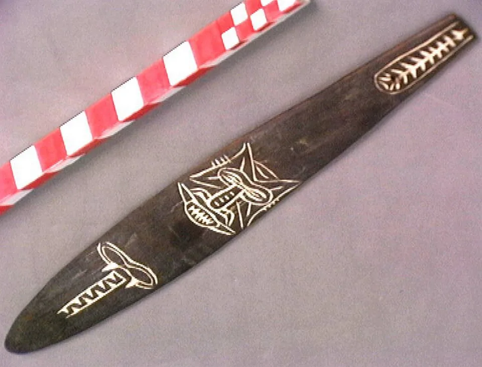 A dark wooden bullroarer blade incised with white-filled figures of a face and a fish-bone motif, beside a red and white scale bar.